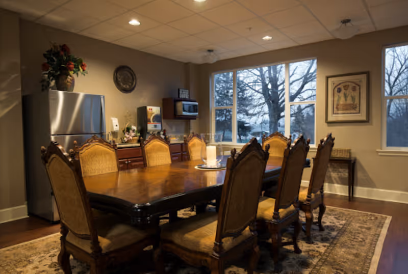 A warmly lit dining room with a large wooden dining table surrounded by eight upholstered chairs. The room features a patterned area rug, a stainless steel refrigerator, a microwave mounted on the wall, a coffee machine, and decorative items including a vase with flowers and framed artwork. Large windows reveal a view of leafless trees outside.