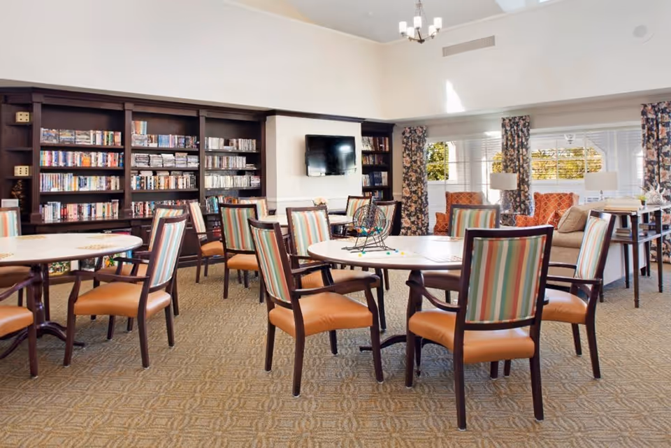 A bright and spacious common area with round tables and chairs featuring striped upholstery. The room includes a large bookshelf filled with books and games, a wall-mounted TV, and comfortable seating with patterned curtains and large windows letting in natural light.