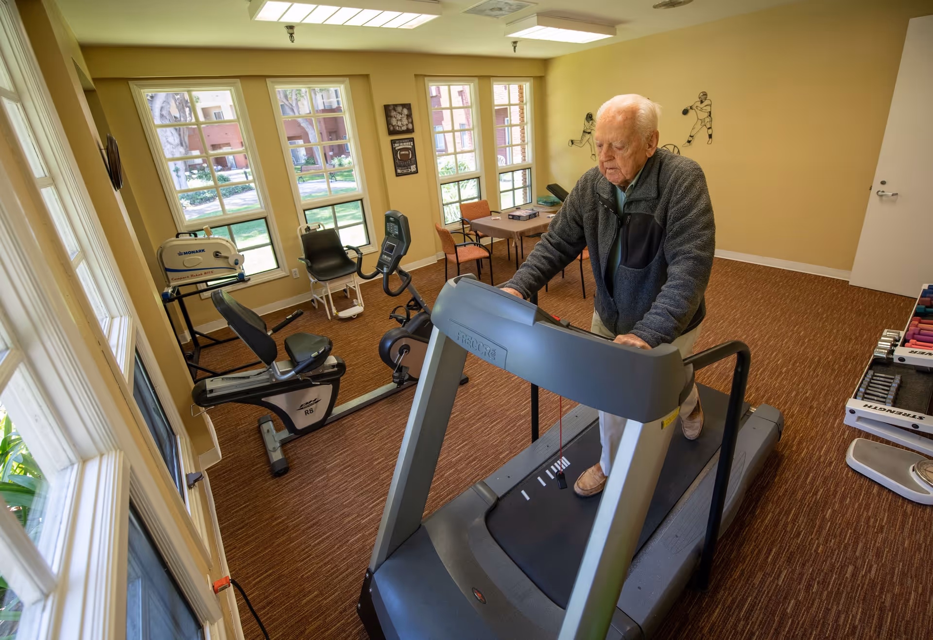 An elderly man walking on a treadmill in a well-lit exercise room with large windows. The room contains various exercise equipment including a recumbent bike, weights, and a table with chairs. The walls are decorated with sports-themed artwork.