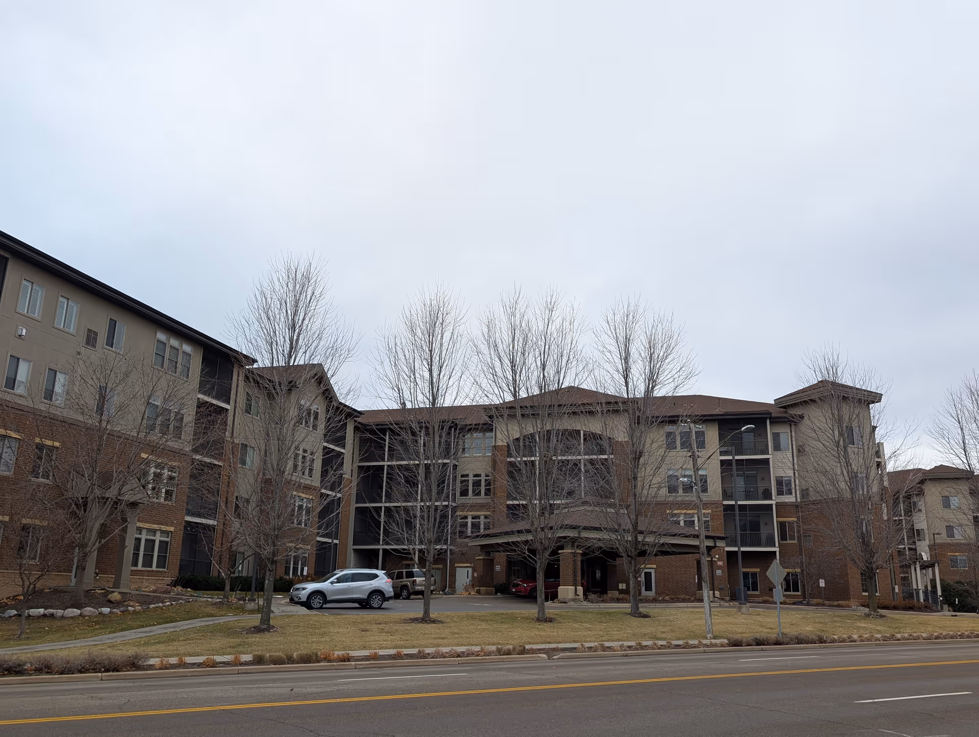 Exterior view of a multi-story senior living facility building with a covered entrance, several leafless trees in front, parked cars, and a road in the foreground under a cloudy sky.