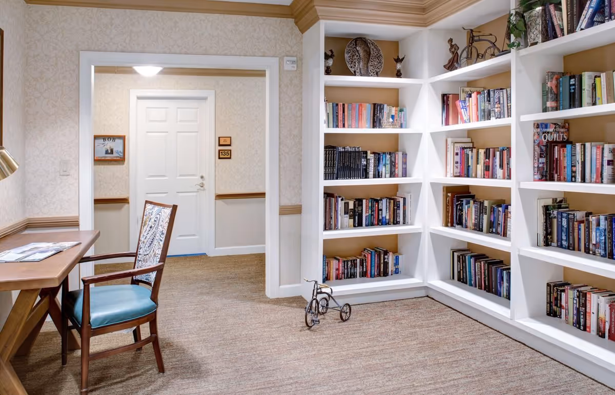 A cozy reading nook in a senior living facility featuring white built-in bookshelves filled with books, a small decorative tricycle on the floor, a wooden desk with a chair upholstered in blue and patterned fabric, and a hallway with a white door labeled 135.