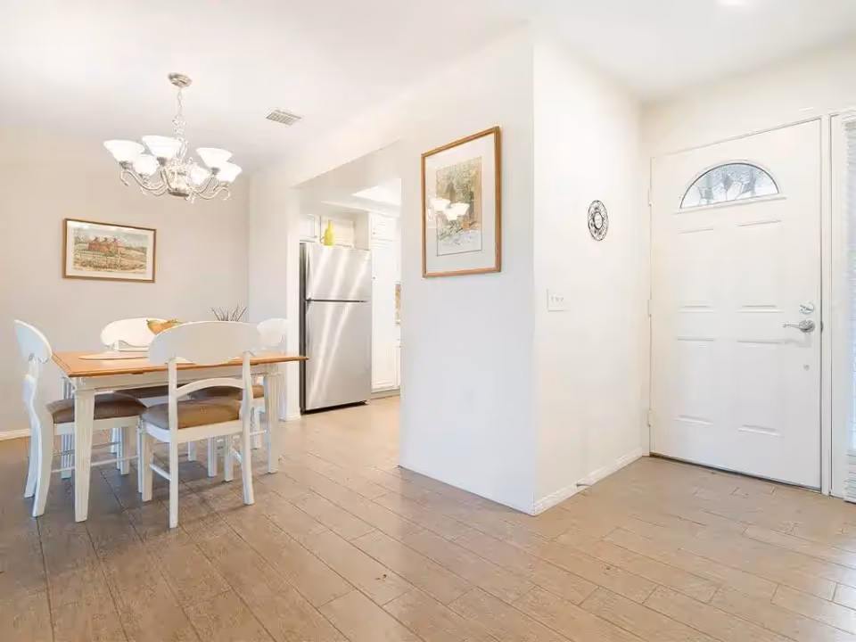 Interior view of a dining area with a wooden table and four white chairs, a chandelier overhead, a framed picture on the wall, and a white front door to the right. A stainless steel refrigerator is visible in the adjacent kitchen area.