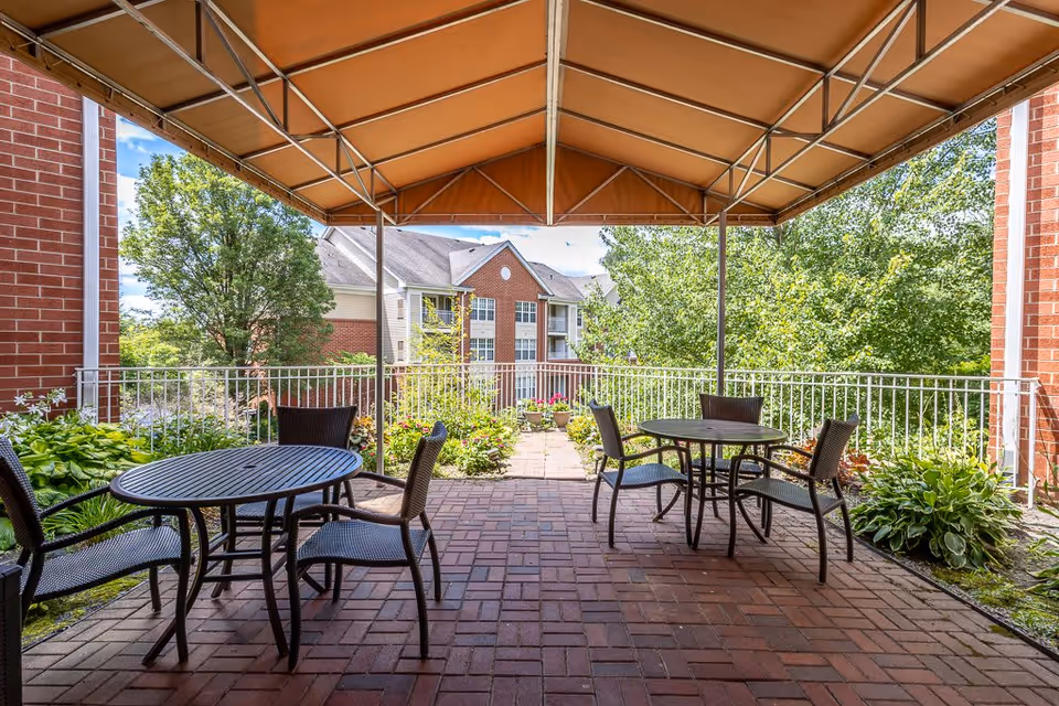 Outdoor covered patio area with two round black metal tables and six matching chairs on a brick floor. The patio is surrounded by greenery including bushes and trees, with a multi-story brick and siding building visible in the background under a partly cloudy sky.
