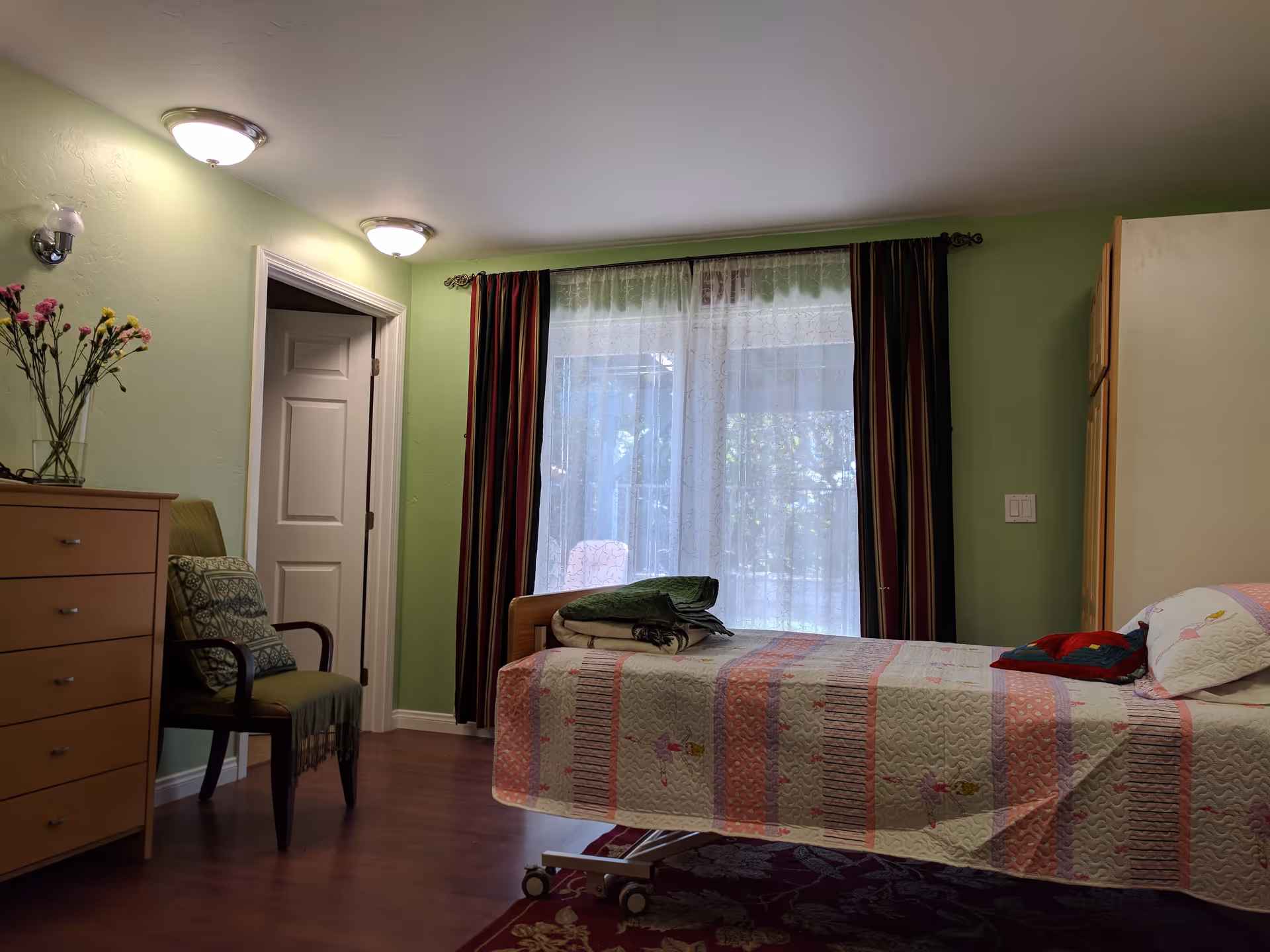 A bedroom with light green walls, a bed with a colorful quilt and pillows, a wooden dresser with a vase of flowers, a chair with cushions, and a large window with sheer and dark curtains letting in natural light.