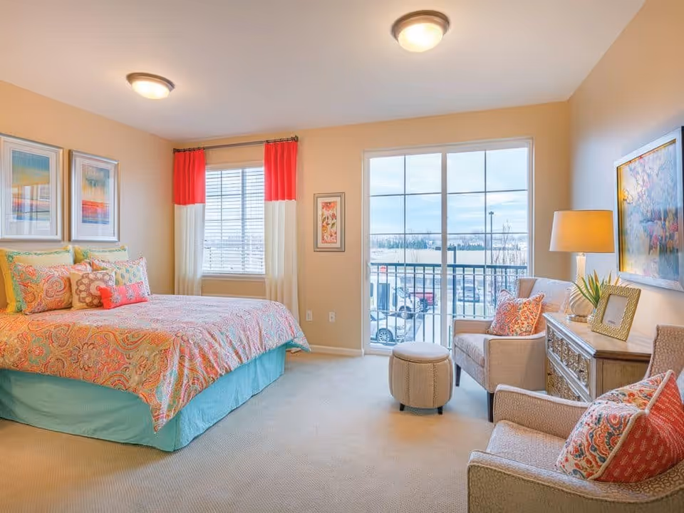 A bright and cozy bedroom featuring a large bed with colorful patterned bedding and multiple pillows. The room has beige walls, two framed artworks above the bed, and a window with red and white curtains. There are two armchairs with patterned cushions, a small round ottoman, a wooden dresser with a lamp, a plant, and a framed photo. A large sliding glass door opens to a balcony overlooking a parking lot and open sky.