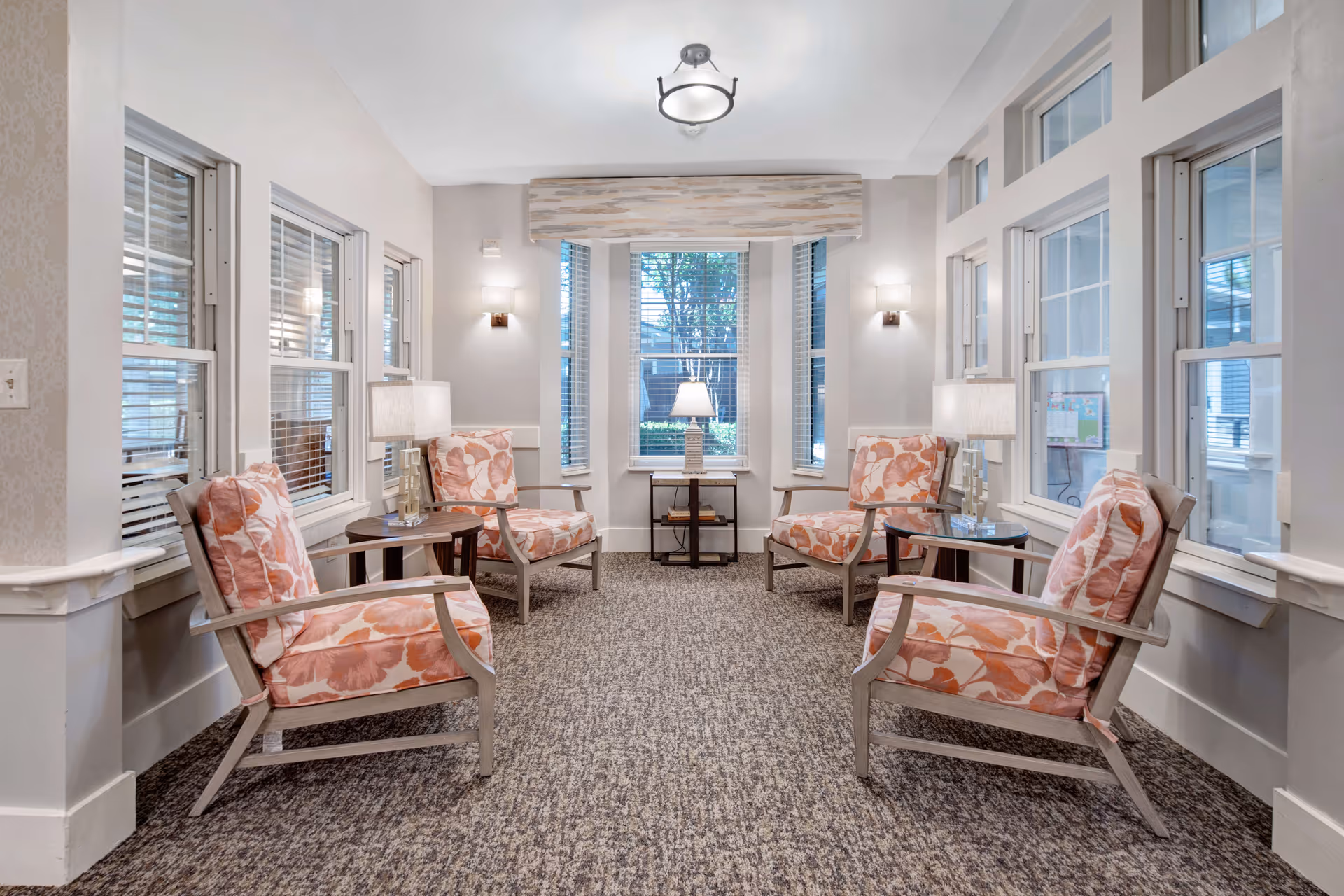 A bright and cozy sitting area with four cushioned armchairs featuring orange floral patterns arranged around two small round tables with lamps. The room has multiple windows with white blinds, light gray walls, and a carpeted floor. A small table with a lamp is positioned in front of a large window at the far end of the room.