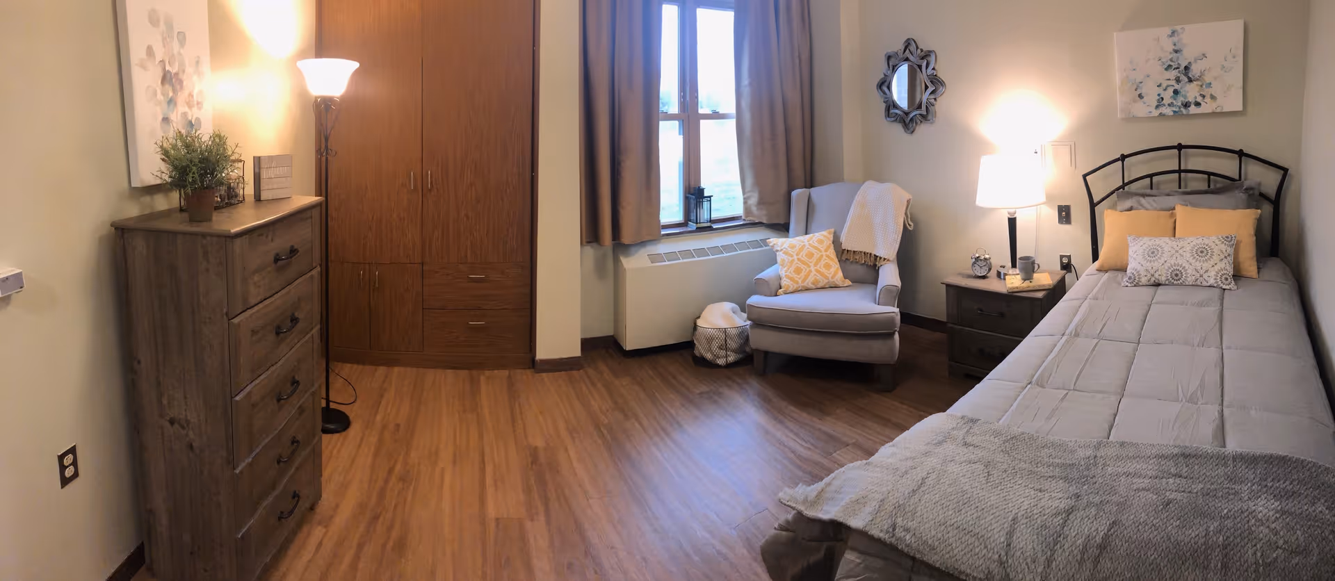 A cozy senior living bedroom with a single bed made with gray bedding and yellow accent pillows. Next to the bed is a wooden nightstand with a lamp, an alarm clock, and a mug. There is a comfortable armchair with a yellow pillow and a blanket near a window with brown curtains. A wooden dresser and a tall wooden wardrobe are also visible, along with a floor lamp and wall art.