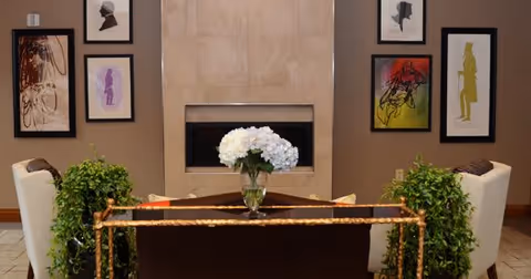 A cozy interior seating area with two white armchairs facing a modern rectangular fireplace set in a tiled wall. A dark wooden coffee table with a glass vase of white flowers is centered between the chairs. Green potted plants flank the seating area, and colorful framed abstract artwork is displayed on the walls.