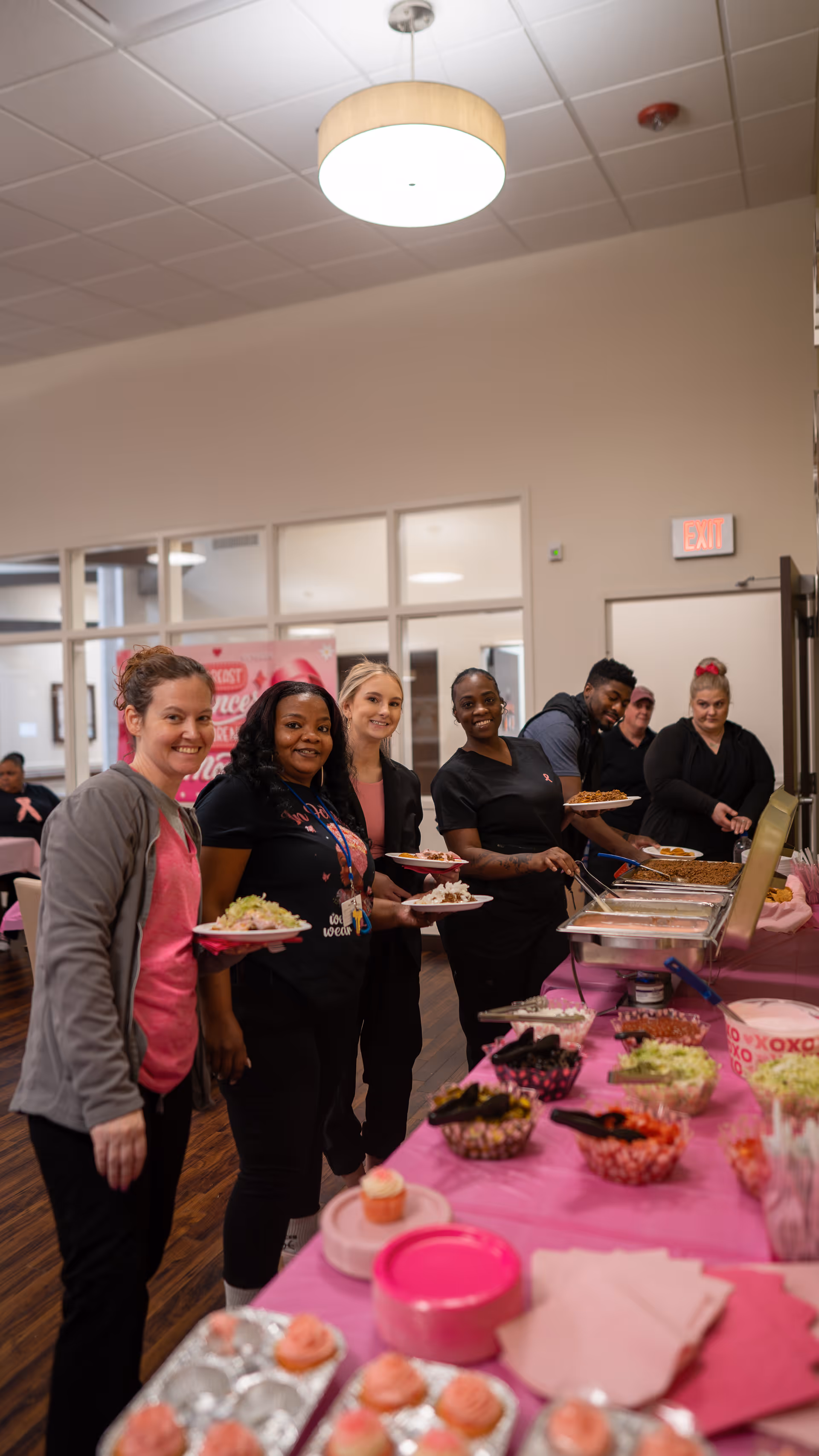 A group of six people standing in line at a buffet table covered with a pink tablecloth, serving themselves food. The setting appears to be an indoor dining or common area with large windows and a ceiling light fixture. The people are smiling and holding plates with food.