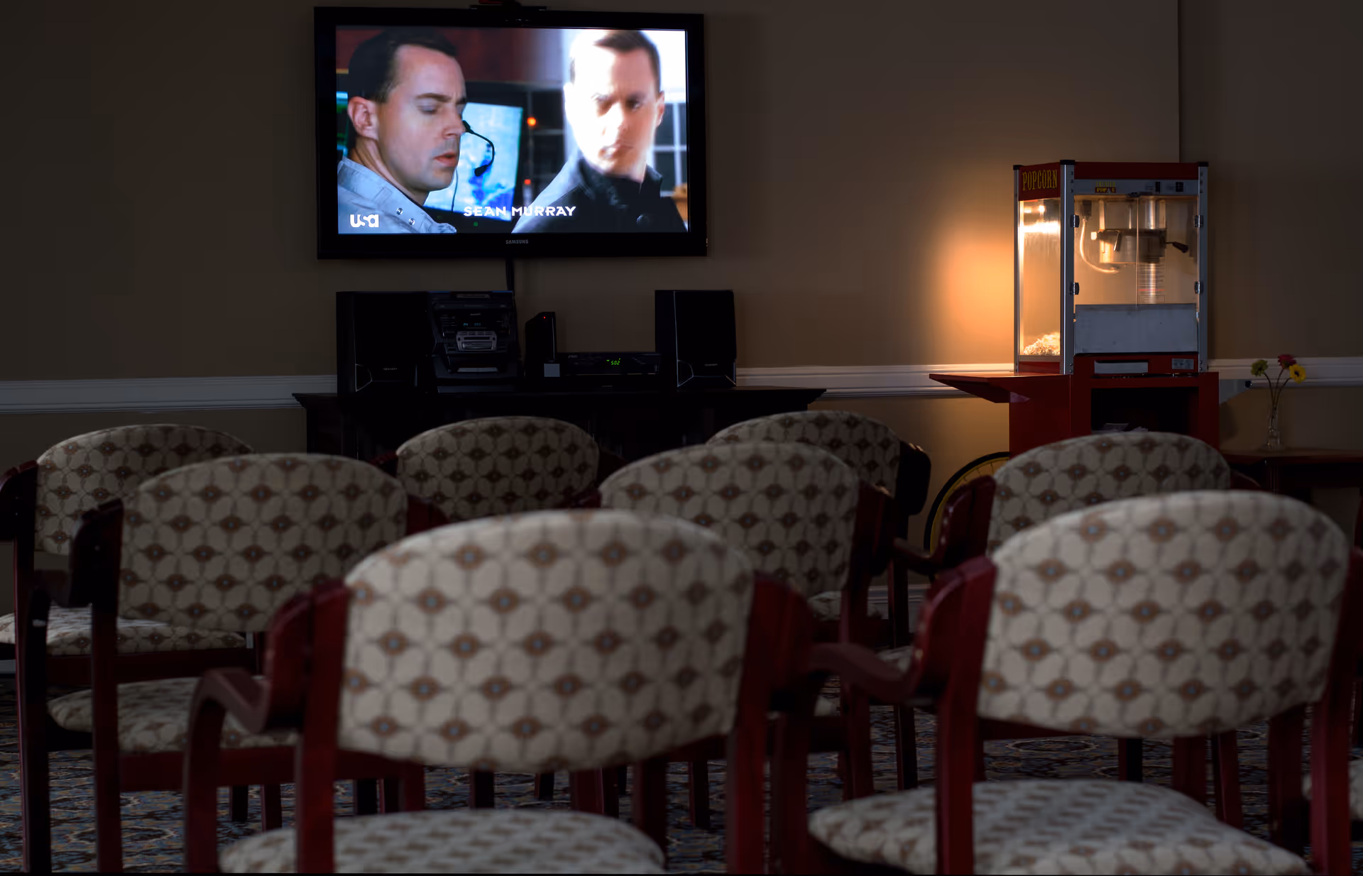 Small screening room with rows of patterned chairs facing a wall-mounted TV and a popcorn machine.