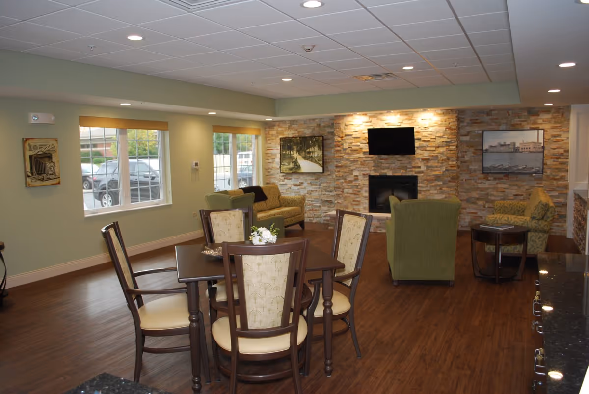 Bright senior living common room with a dining table and chairs, upholstered seating, a TV above a stone fireplace, and large windows.