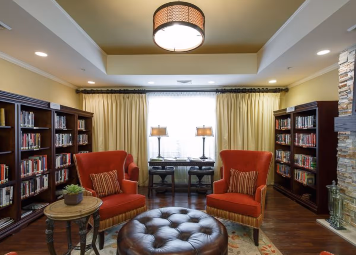 Cozy interior reading room with bookshelves, two red armchairs, a tufted leather ottoman, table lamps in front of a curtained window, and a stone fireplace.