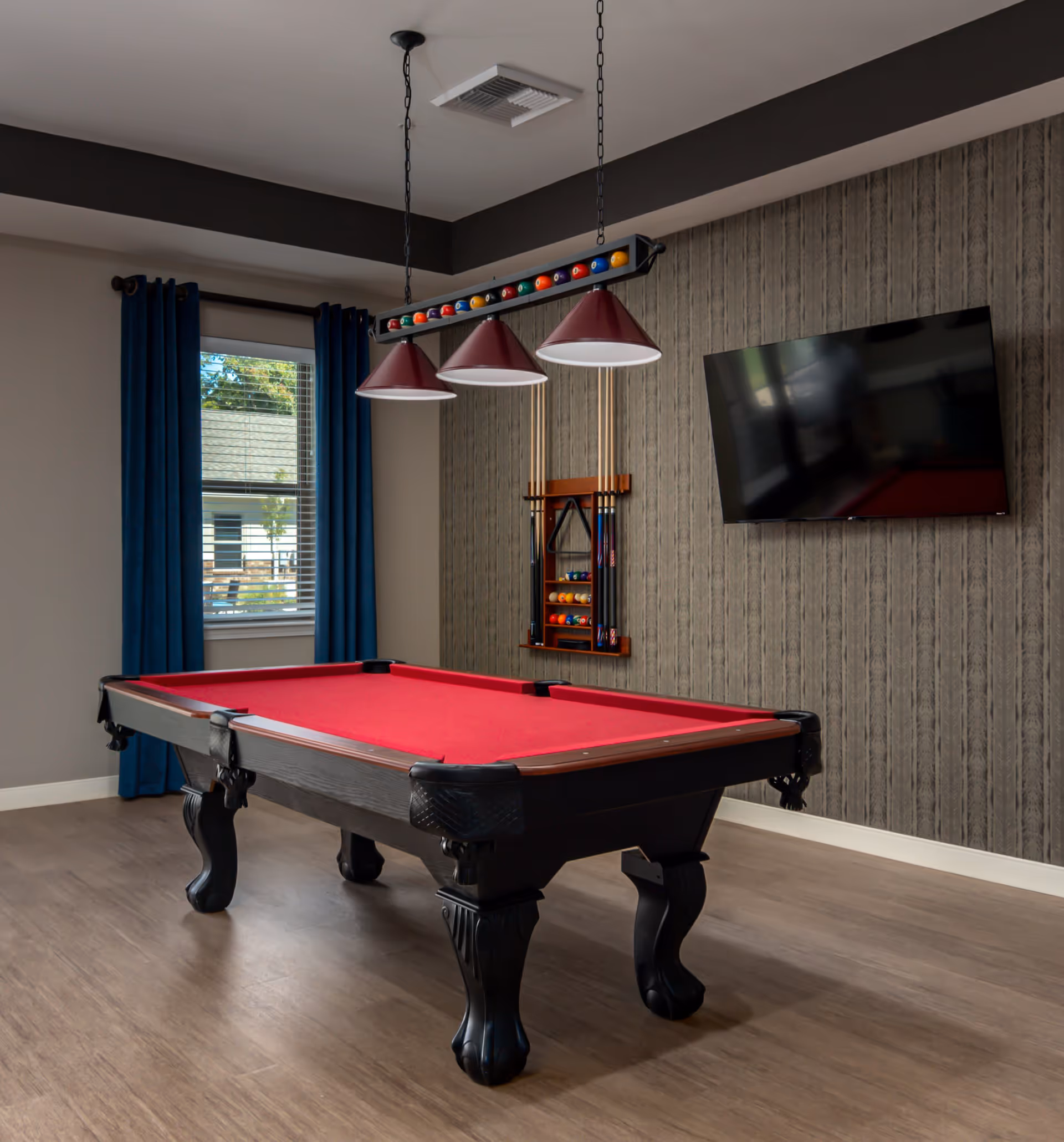 A game room with a red felt pool table, three hanging pendant lights above it with a rack of pool cues and balls mounted on a textured gray wall. A flat-screen TV is mounted on the wall to the right, and a window with blue curtains is on the left side of the room.