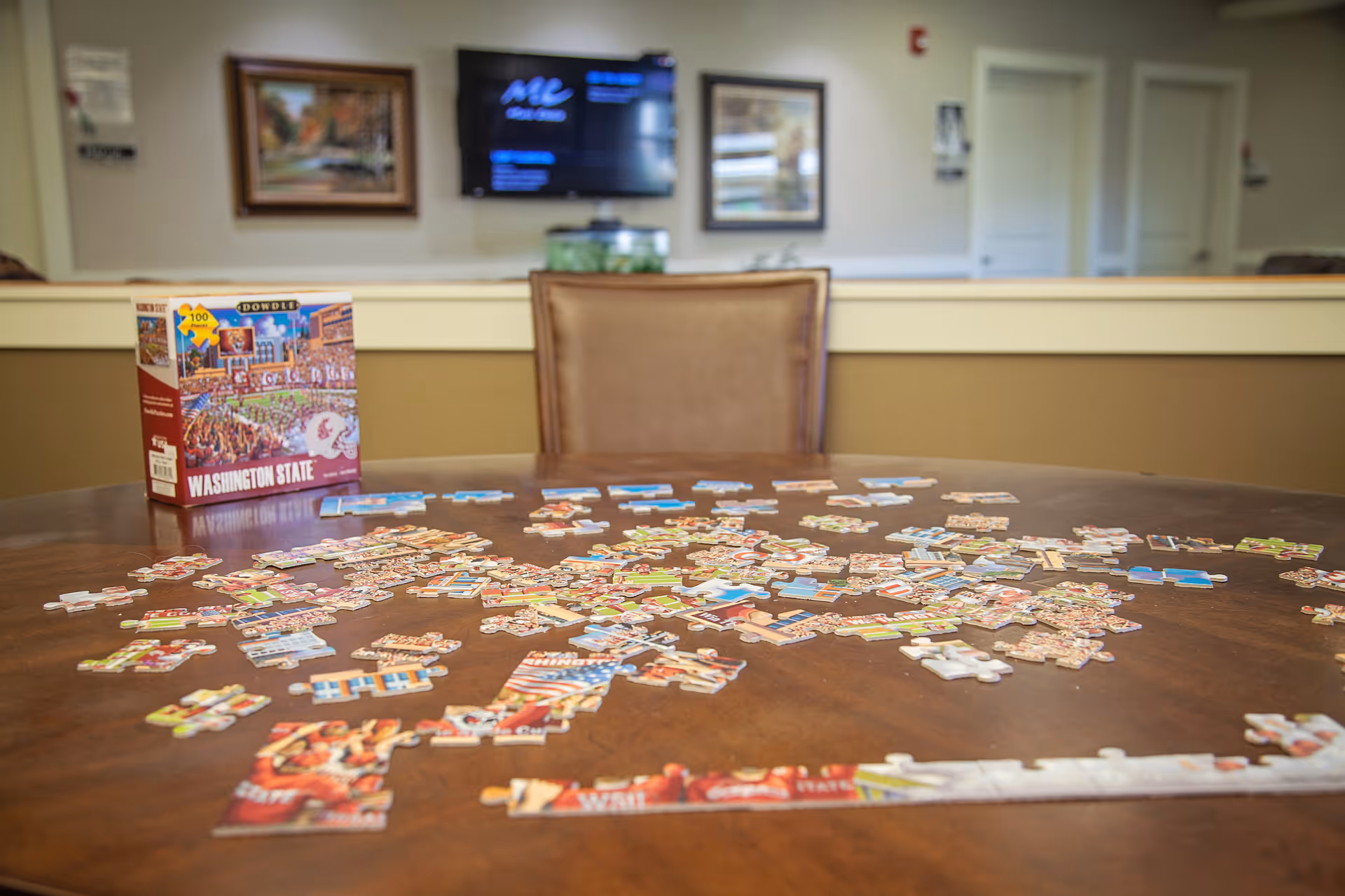 A wooden table with scattered puzzle pieces and a puzzle box labeled 'Washington State' on it. In the background, there is a brown chair, a wall with framed pictures, and a television mounted on the wall.