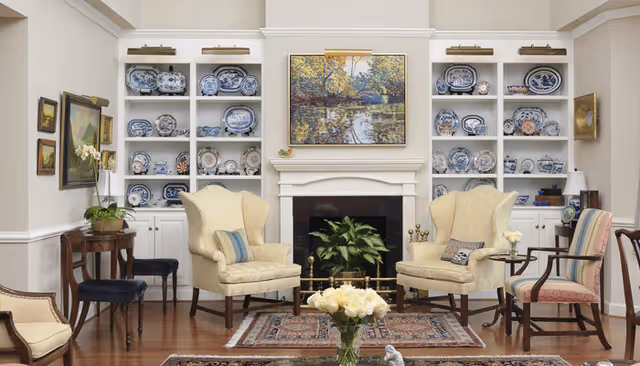 A cozy living room with two cream-colored wingback chairs and a striped armchair arranged around a fireplace. The fireplace has a plant inside and a landscape painting above it. Built-in shelves on either side of the fireplace display blue and white decorative plates. There are small tables with lamps and flowers, and a patterned rug on the wooden floor.