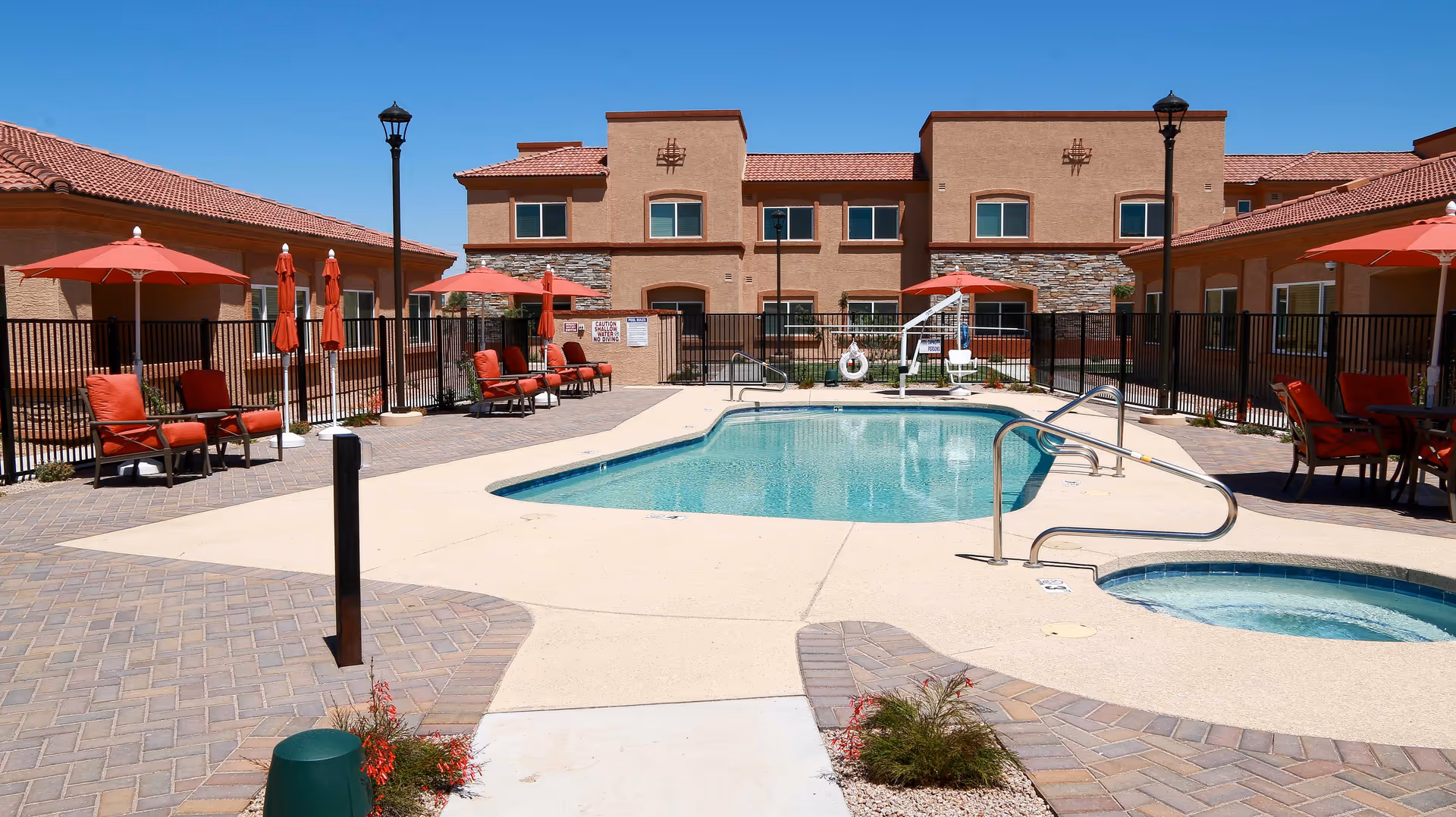 Outdoor swimming pool area at a senior living facility with red cushioned lounge chairs and umbrellas around the pool. The building in the background has a tan stucco exterior with red tile roofing under a clear blue sky.