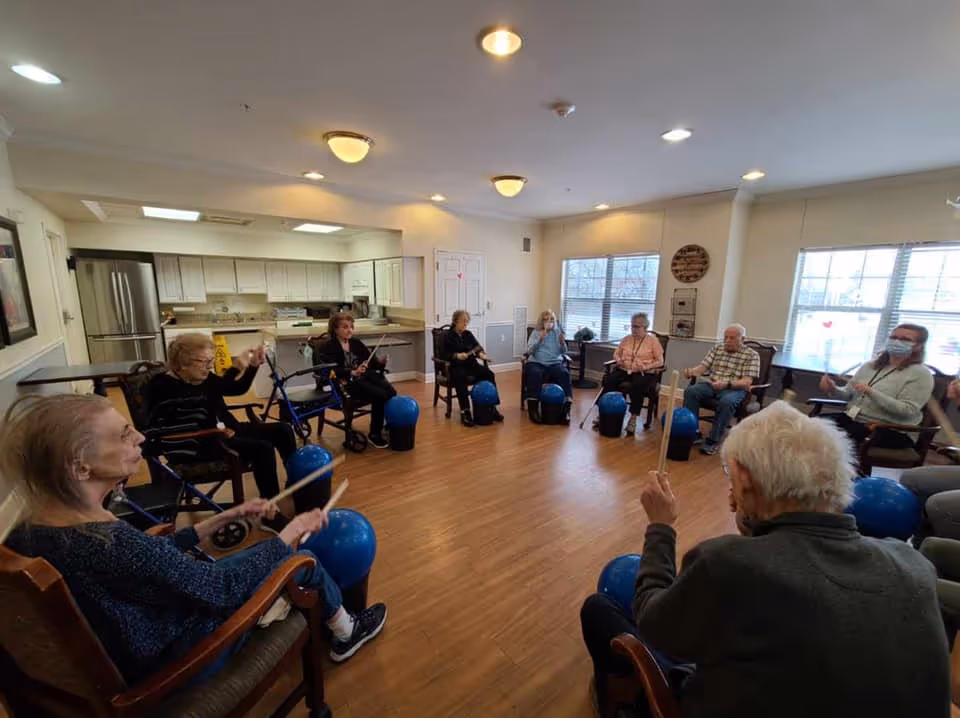 A group of elderly people seated in a circle in a spacious room with wooden flooring and large windows. They are participating in a group activity using blue exercise balls and drumsticks. The room has a kitchen area in the background with white cabinets and stainless steel appliances.