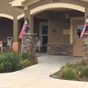 Covered entrance area of a senior living facility with stone pillars, American flags, rocking chairs, and a walkway surrounded by plants and flowers.