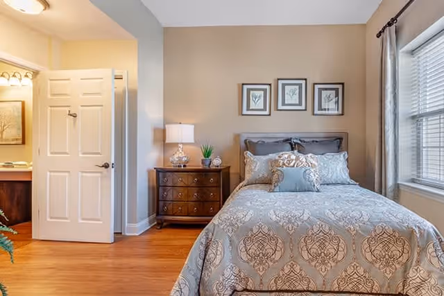 A neatly made bed with patterned bedding in a bedroom. There is a wooden nightstand with a lamp, a small plant, and a clock next to the bed. Three framed botanical prints hang on the wall above the bed. A window with blinds and curtains is on the right side, and an open door leads to a bathroom area with a sink and mirror visible.