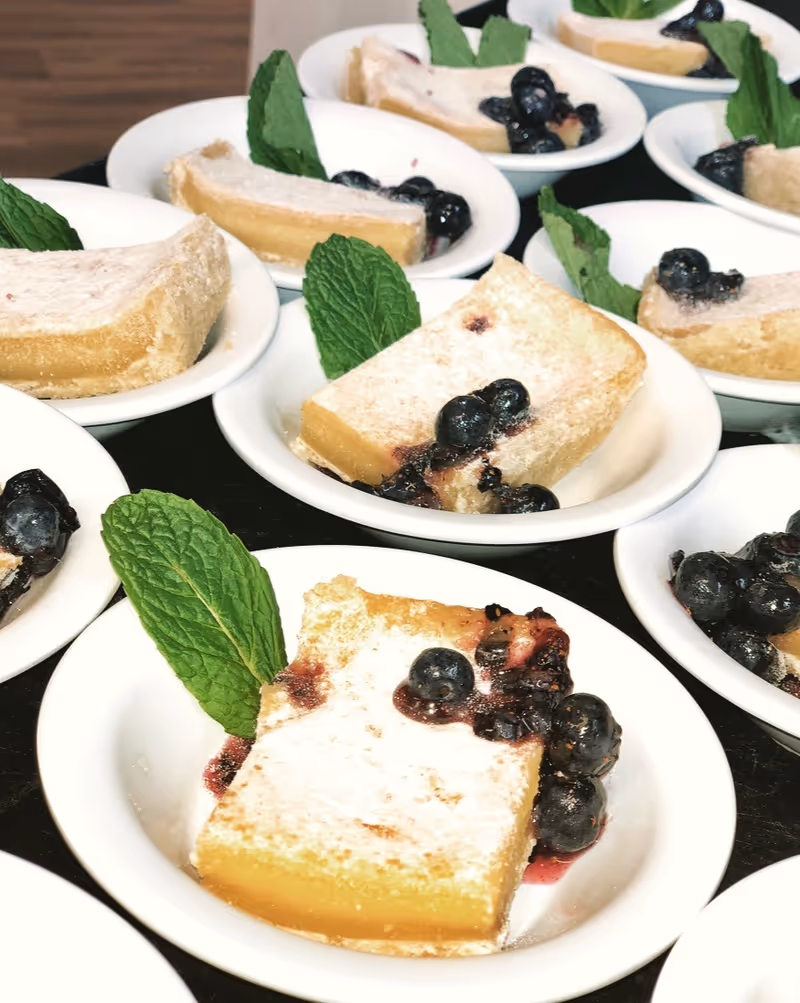 Small white dishes each holding a powdered lemon bar garnished with blueberries and a mint leaf.