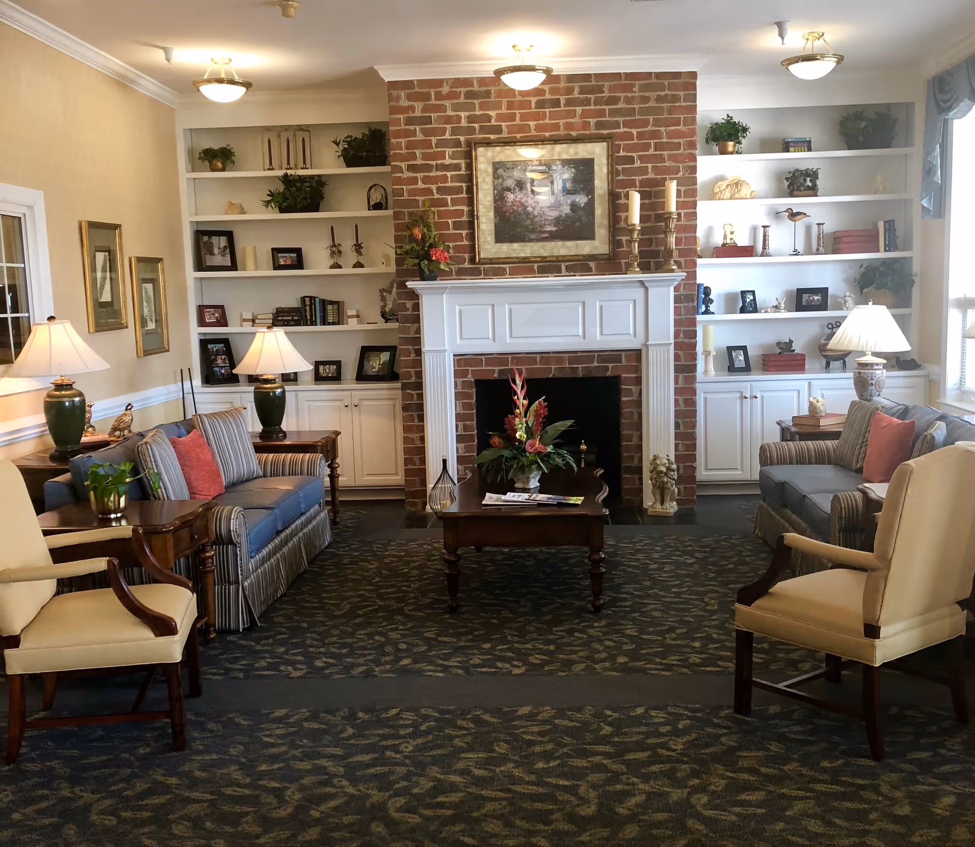 A cozy living room with two striped sofas and two beige armchairs arranged around a wooden coffee table with a floral centerpiece. The room features a brick fireplace with a white mantel, decorated with candles and a framed painting above it. Built-in white shelves on either side of the fireplace hold various decorative items and framed photos. The room is softly lit by table lamps and ceiling lights, with a patterned carpet covering the floor.