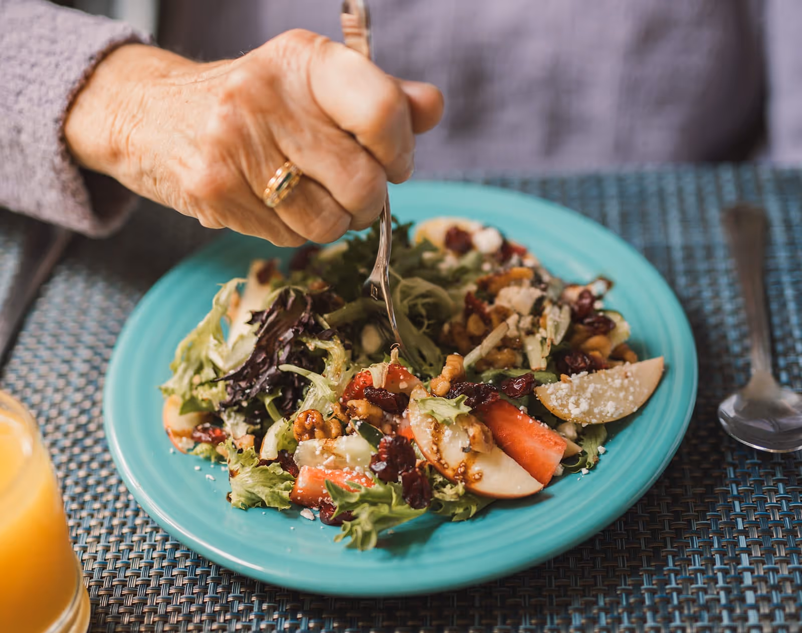 An elderly person's hand holding a fork over a plate of salad with a glass of orange juice nearby.