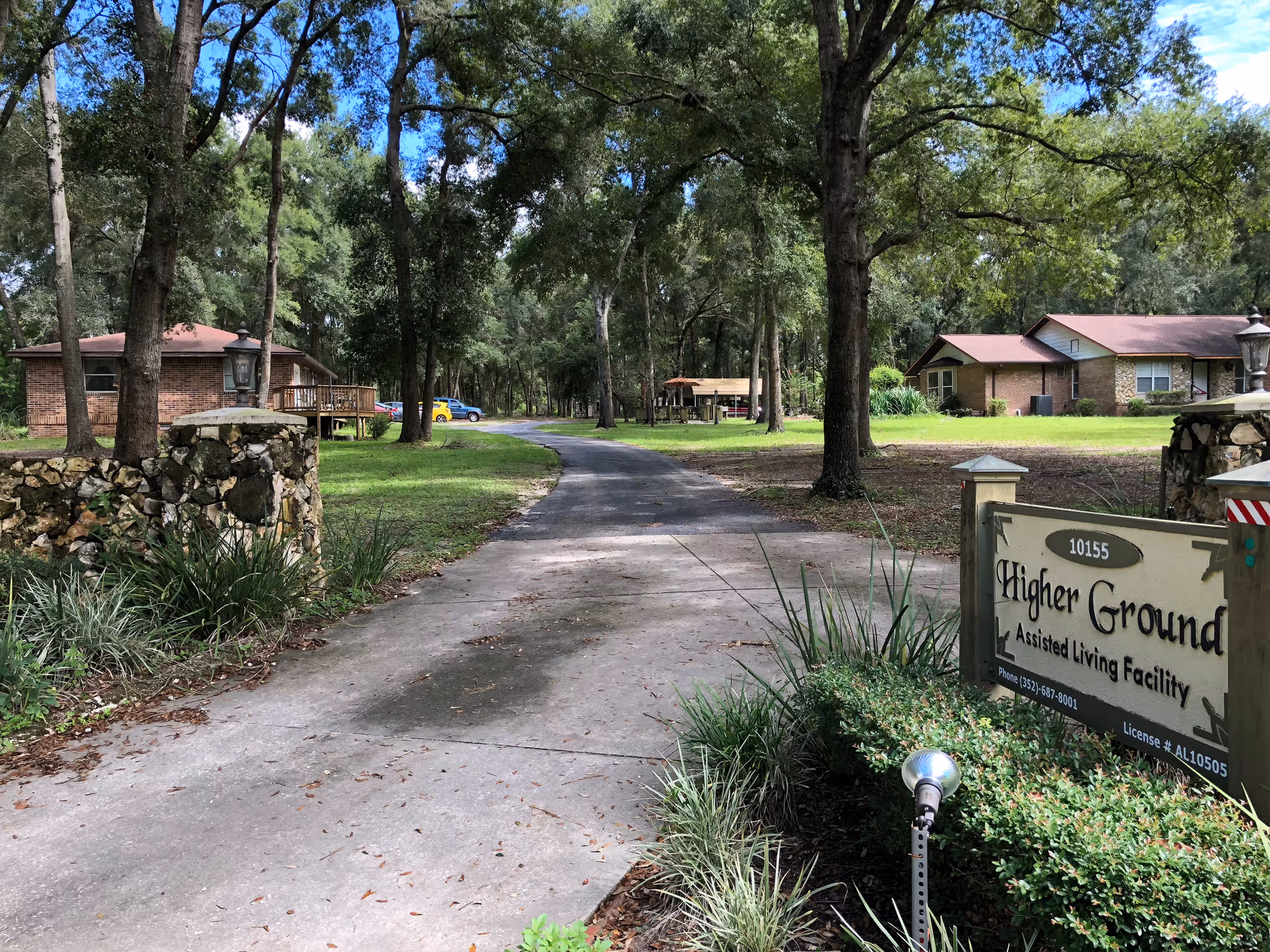 A paved driveway leading into a wooded area with tall trees and green grass on either side. There are two single-story brick buildings with brown roofs visible on the left and right sides. A sign near the entrance reads 'Higher Ground Assisted Living Facility' with contact information. The sky is clear and blue.