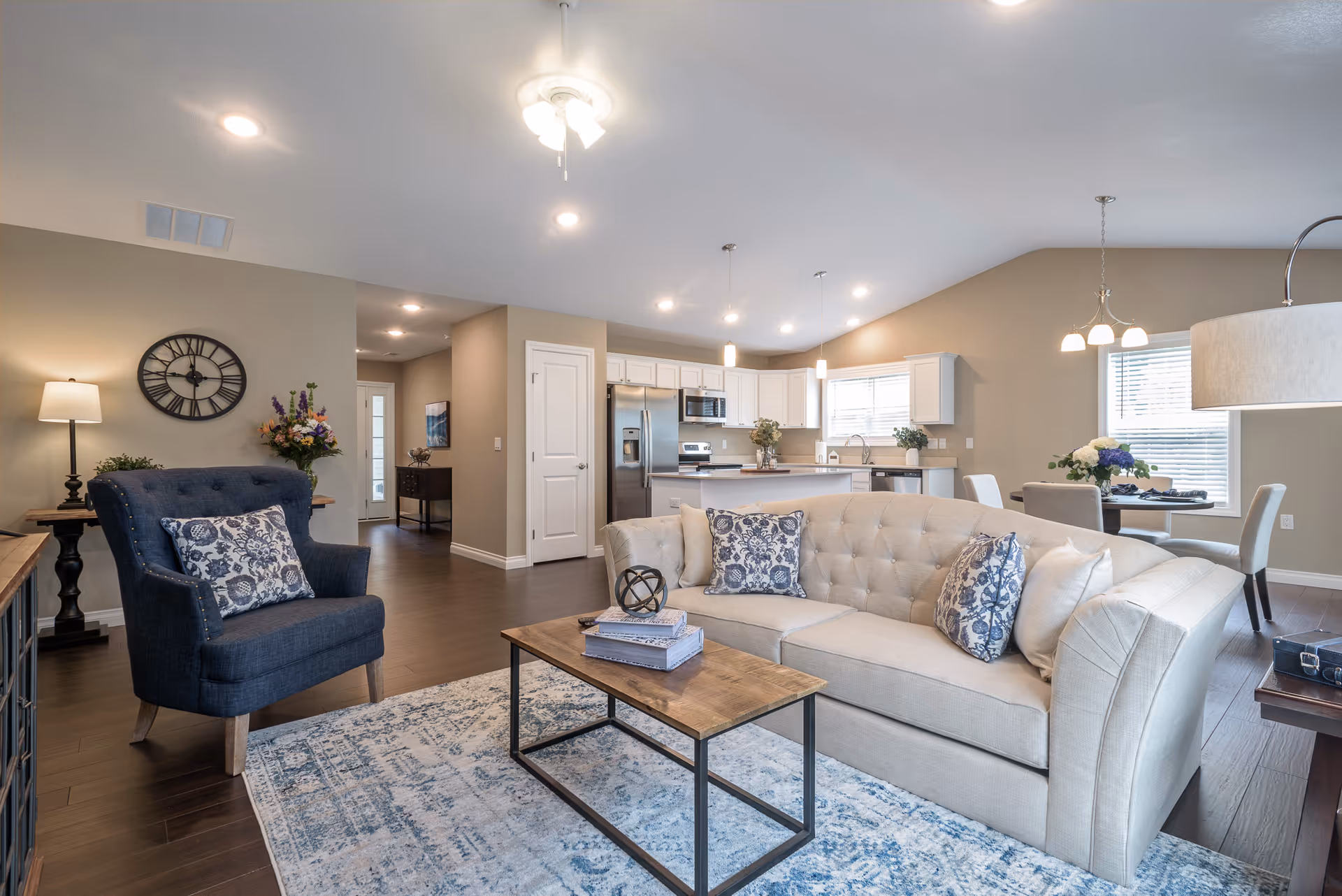 A spacious and well-lit living area featuring a beige tufted sofa with patterned cushions, a wooden coffee table with decorative items, and a dark blue armchair with a floral cushion. The room has wooden flooring, a large area rug, and a modern ceiling fan with lights. In the background, there is an open kitchen with white cabinets, stainless steel appliances, and a dining area with a table and chairs near a window.