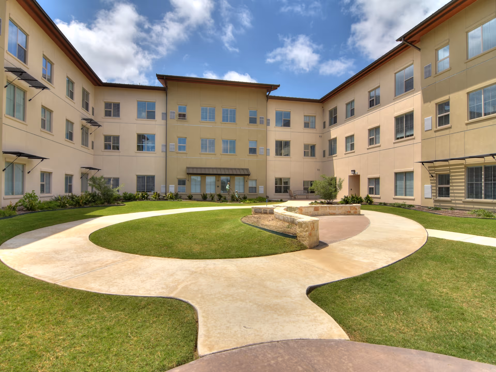 Outdoor courtyard area of a senior living facility with a circular concrete walkway surrounding a grassy lawn and a stone bench. The courtyard is enclosed by a three-story beige building with multiple windows and a partly cloudy blue sky overhead.