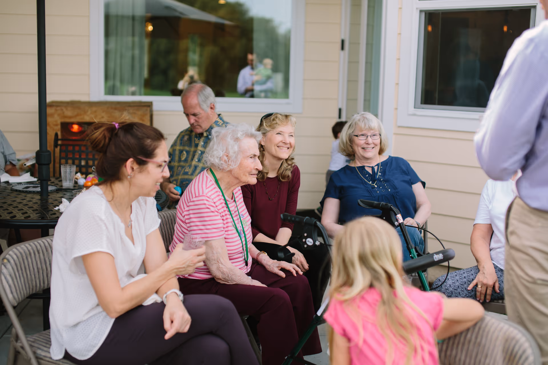 A group of elderly and middle-aged people sitting and smiling together outdoors near a building with beige siding and white-framed windows. Some are seated on folding chairs, and one elderly woman has a walker. A young girl with blonde hair is in the foreground, facing away from the camera.