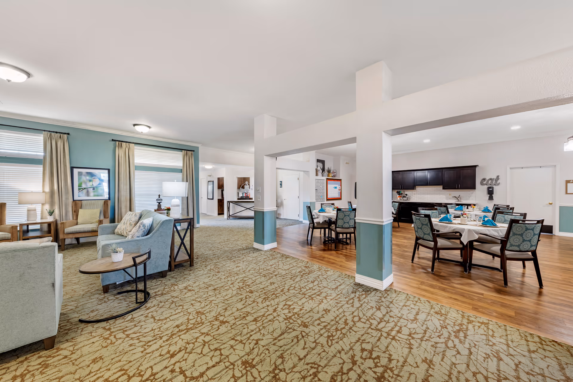 Bright communal lounge and dining area in a senior living facility with seating, tables set for meals, and an open kitchenette.