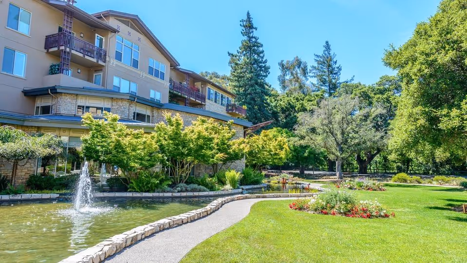 Exterior view of a multi-story senior living building with a landscaped pond, fountain, walkway, and trees.