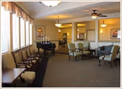 A spacious and well-lit common area with multiple chairs arranged around small tables and a black piano near large windows with blinds. The room features carpeted flooring, ceiling fans with lights, framed artwork on the walls, and a warm, inviting atmosphere.