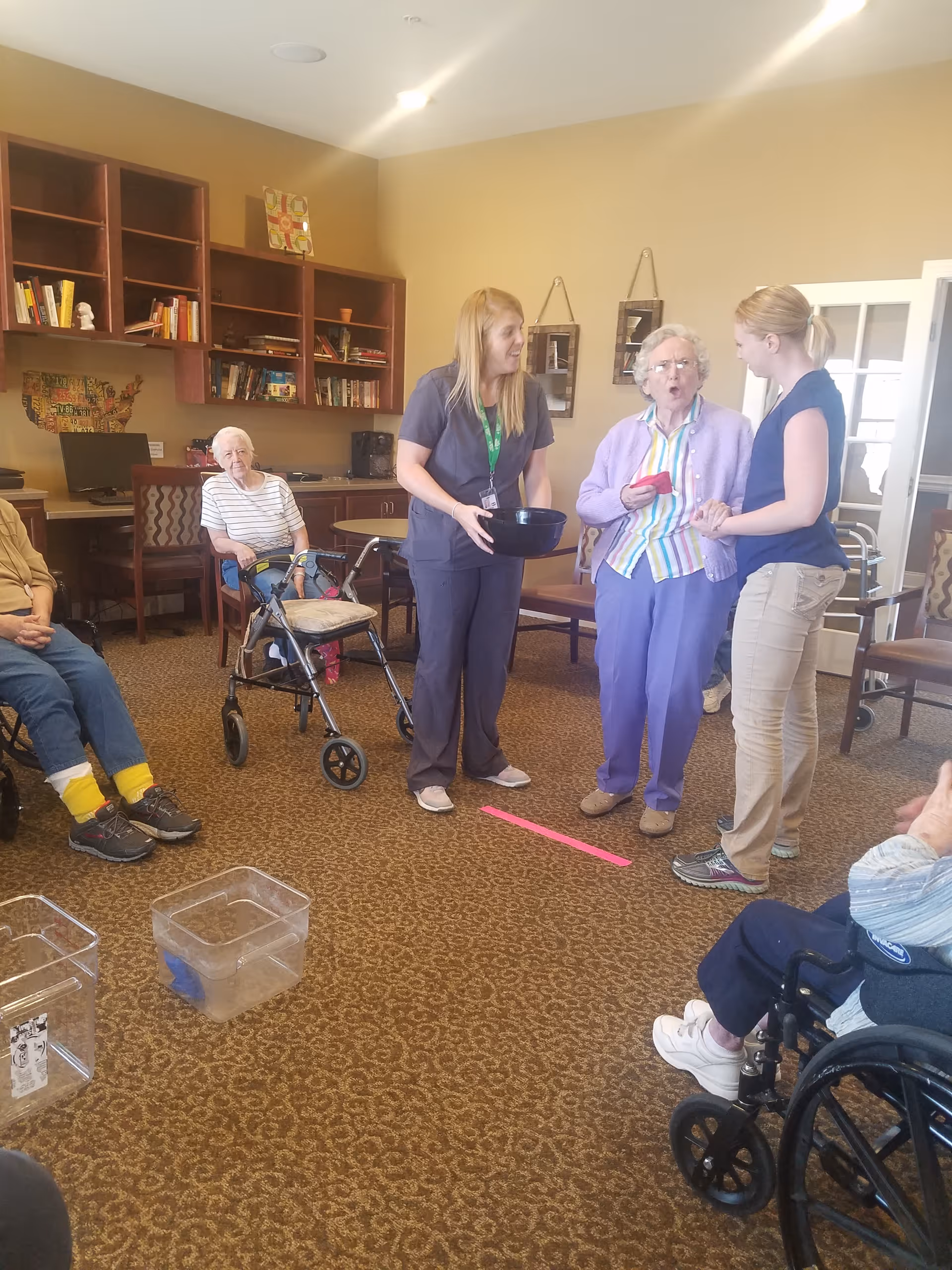 A group of elderly people and two caregivers are gathered in a common room with carpeted floor and wooden bookshelves. One elderly woman is standing and holding a red object while interacting with a caregiver. Other elderly individuals are seated, some in wheelchairs and one with a walker. The room has beige walls and framed pictures hanging on the wall.