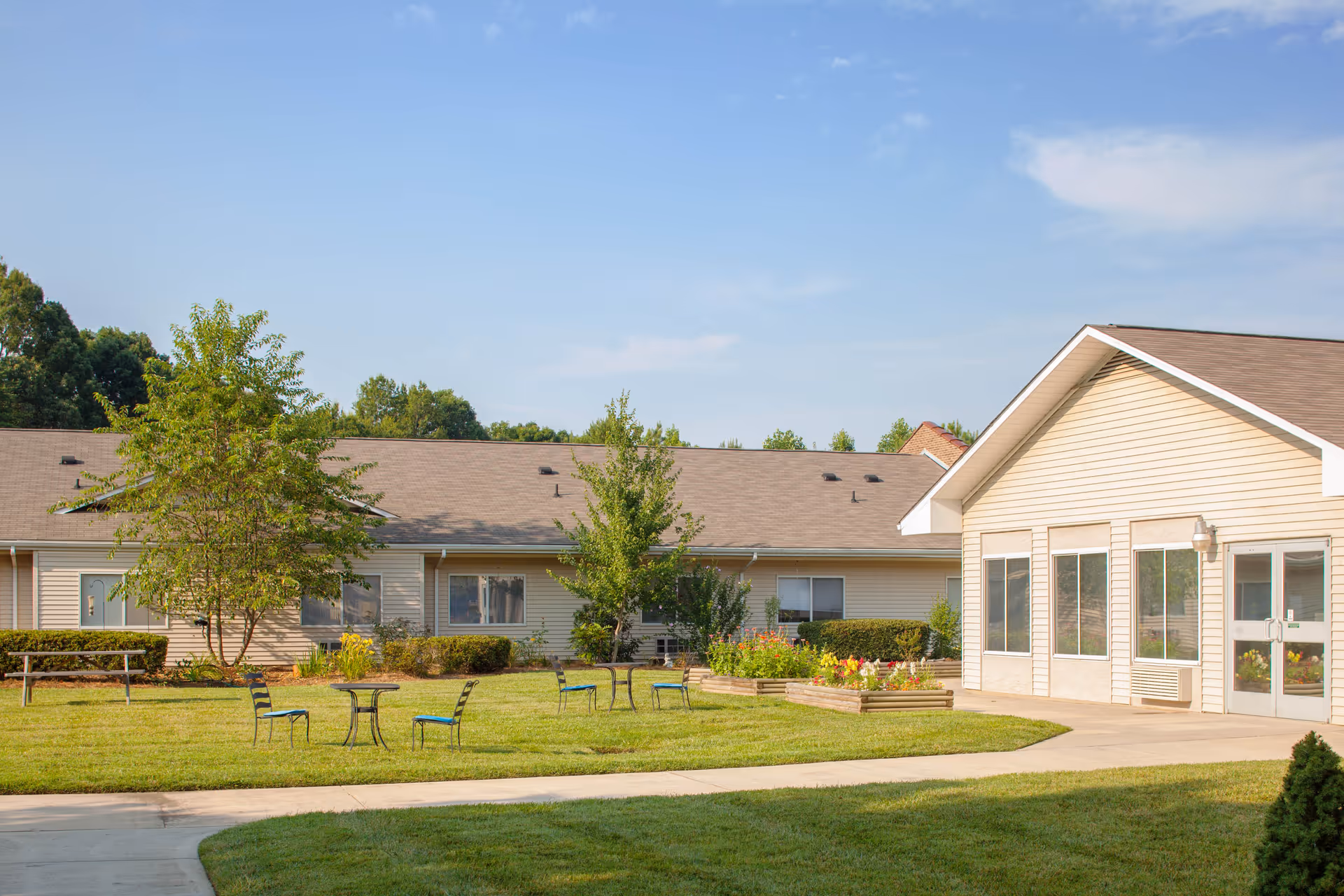 Outdoor view of Mooresville Center showing a well-maintained lawn with several small tables and chairs, a picnic bench, flower beds, and a beige building with multiple windows under a clear blue sky.