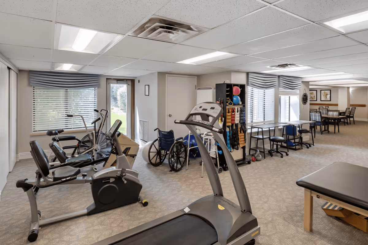 A senior living facility exercise room with various fitness equipment including a treadmill, stationary bikes, and a rowing machine. There is a wheelchair and exercise balls near a storage rack holding resistance bands and other workout accessories. The room has carpeted floors, large windows with blinds, and tables and chairs in the background.