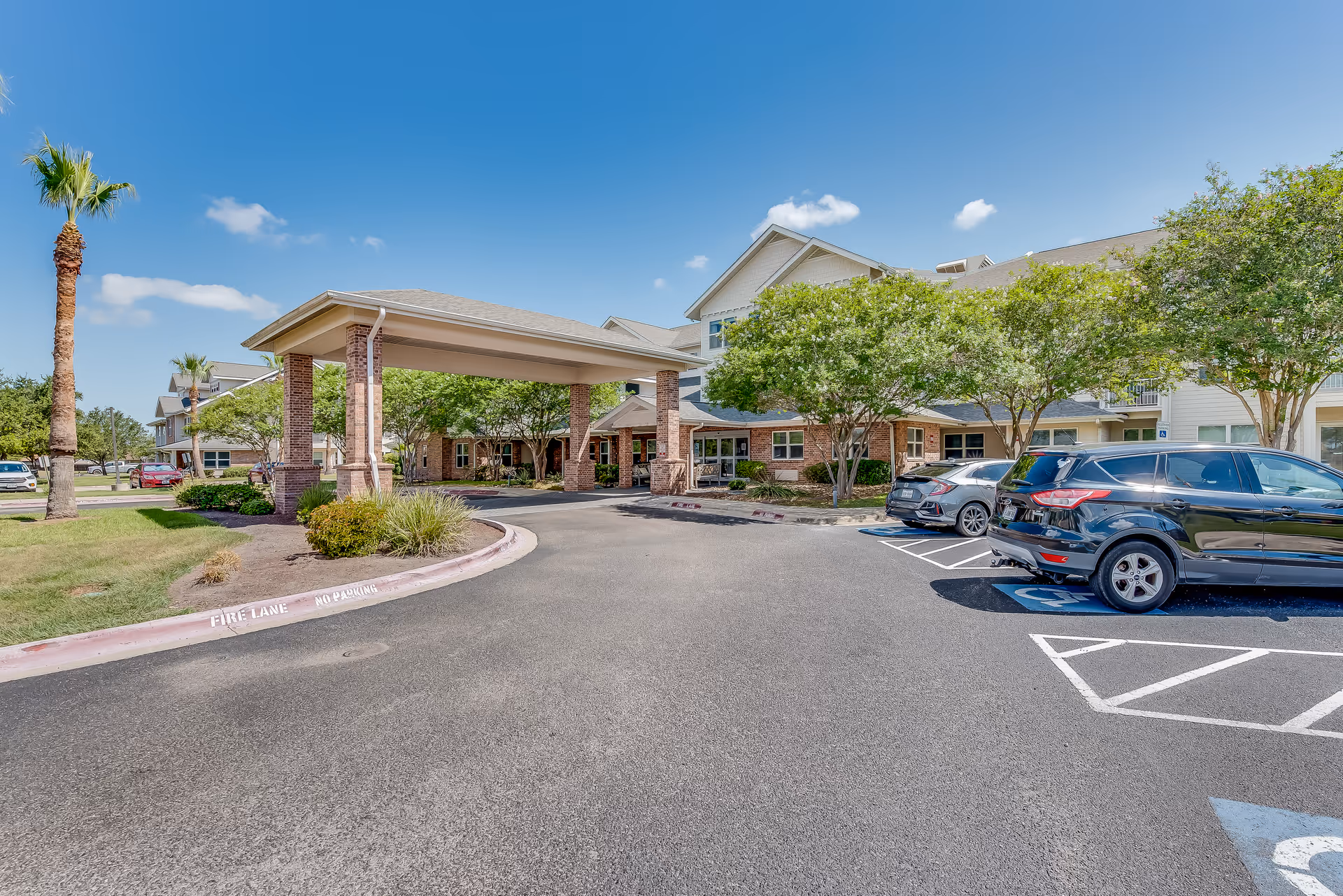 Exterior view of Solstice Senior Living at Corpus Christi showing the entrance with a covered drop-off area, surrounded by trees and parked cars in designated parking spaces under a clear blue sky.