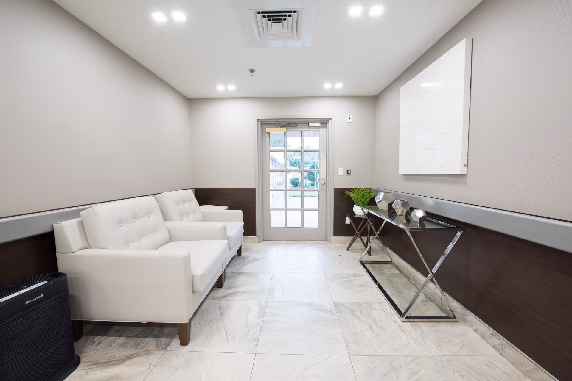 A small waiting area with two white cushioned armchairs on the left side, a glass door at the end, and a modern console table with decorative items and a small plant on the right side. The floor is tiled with light-colored marble, and the walls are painted beige with a dark wood panel halfway up. The ceiling has recessed lighting.