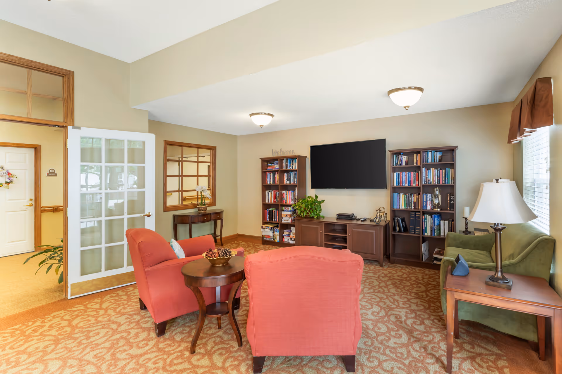 A cozy common sitting room with two red armchairs, a green sofa, bookshelves, a wall-mounted TV, side tables and a glass-paned door.