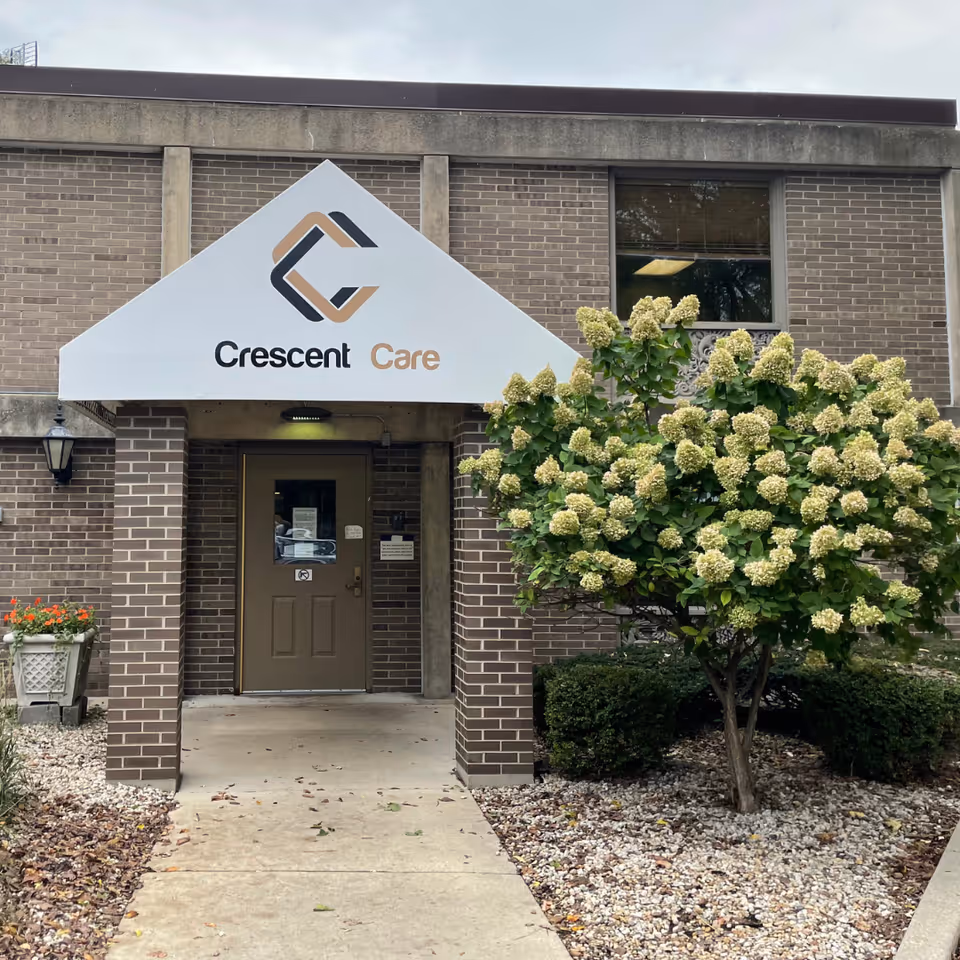 Entrance to Crescent Care facility with a brown door under a white triangular awning displaying the Crescent Care logo. The building is made of brown bricks and there is a flowering shrub with light green flowers to the right of the entrance.