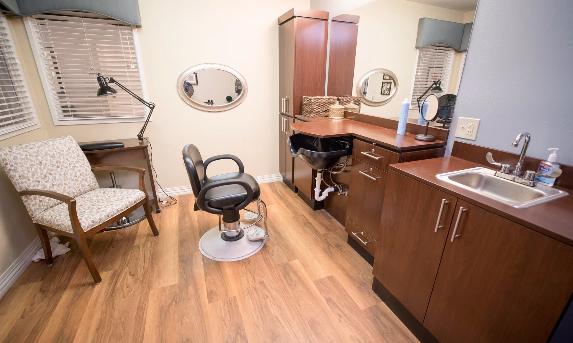 A small salon room with a black salon chair in front of a wooden counter with a sink and a wash basin. There is a floral patterned armchair near the window with blinds, a desk lamp, and two oval mirrors on the wall.