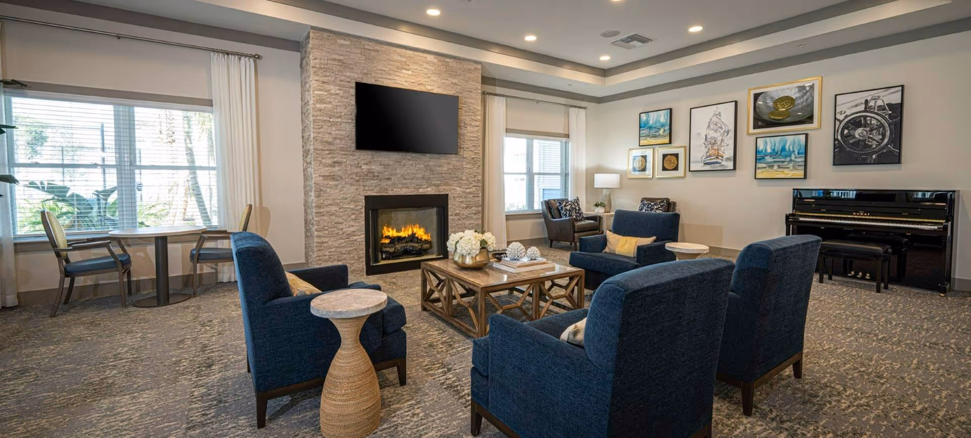 Bright communal lounge with blue armchairs arranged around a coffee table in front of a stone fireplace and wall-mounted TV.