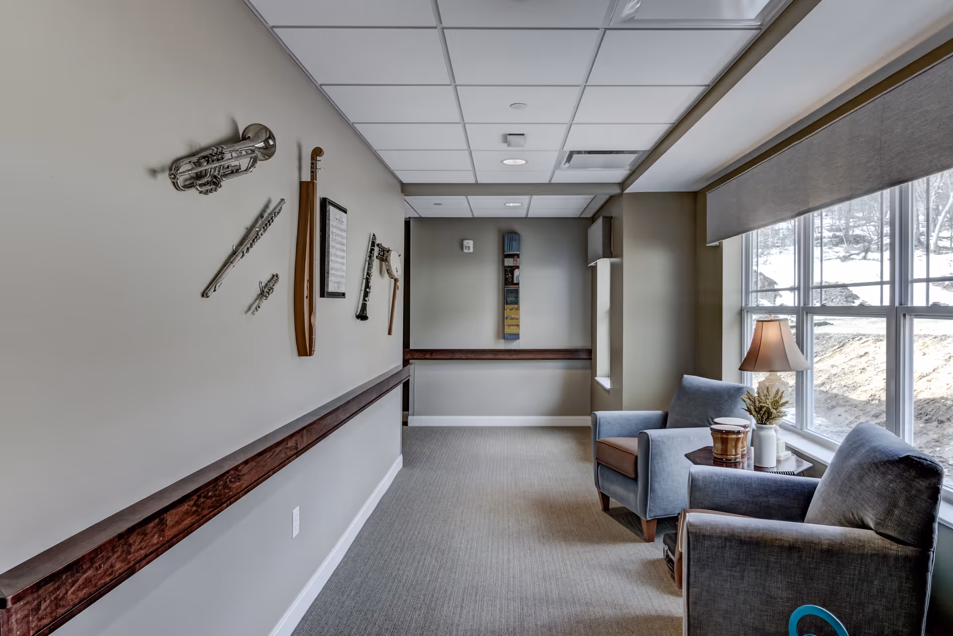 A quiet sitting area in a senior living facility hallway with two blue armchairs next to a window. A small table between the chairs holds a lamp and decorative items. The wall opposite the chairs features musical instruments mounted as decoration, including a trumpet, flute, clarinet, and banjo. The space is carpeted and has a drop ceiling with recessed lighting.