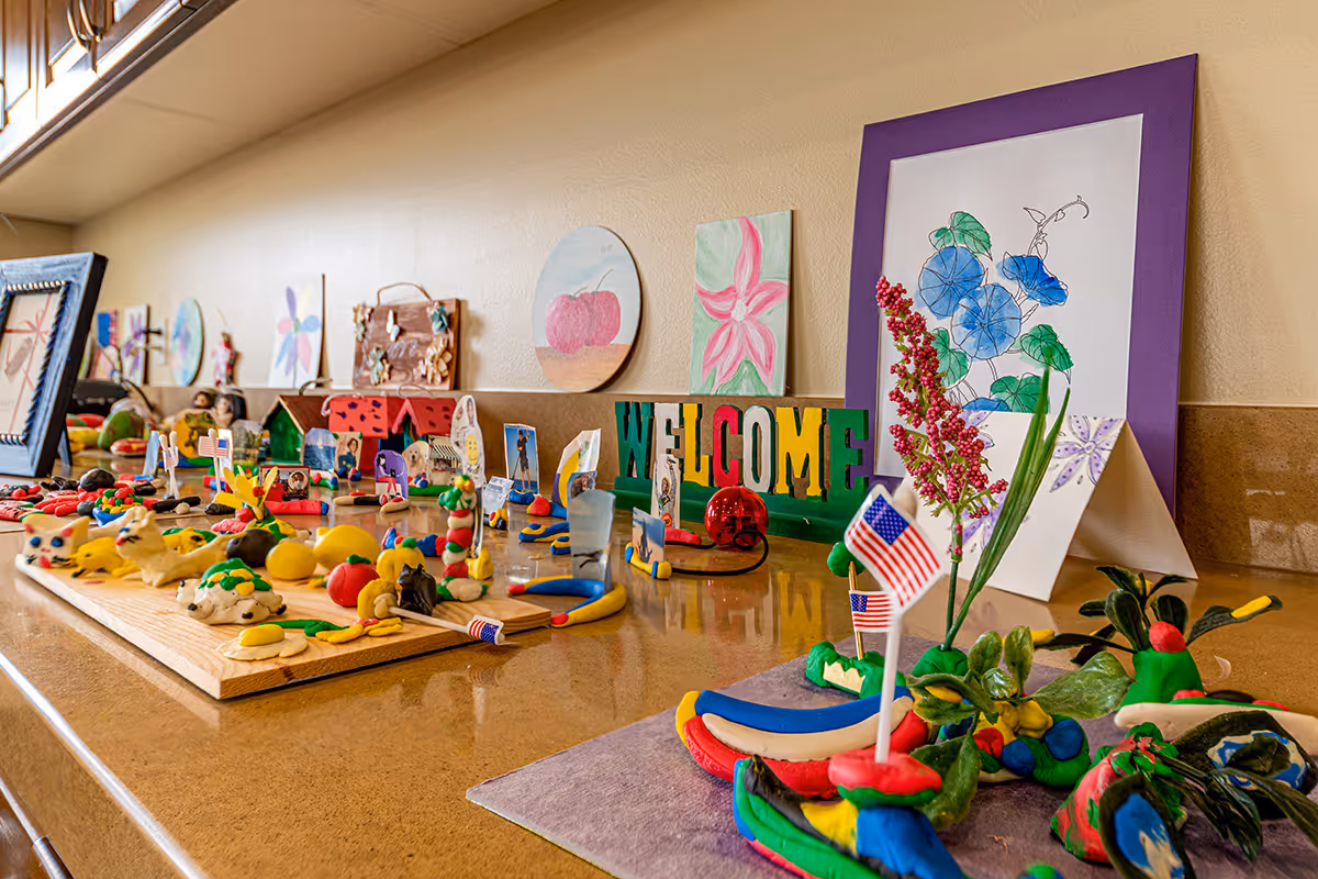A countertop display featuring colorful handmade clay models, small American flags, and various pieces of artwork including paintings of flowers and a 'WELCOME' sign, set against a beige wall.