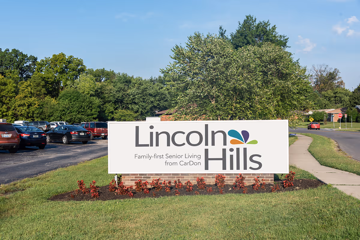 Outdoor view of a large white sign with the text 'Lincoln Hills Family-first Senior Living from CarDon' placed on a brick base with small red plants in front. The sign is located on a grassy area next to a sidewalk and a road, with trees and parked cars visible in the background under a clear blue sky.