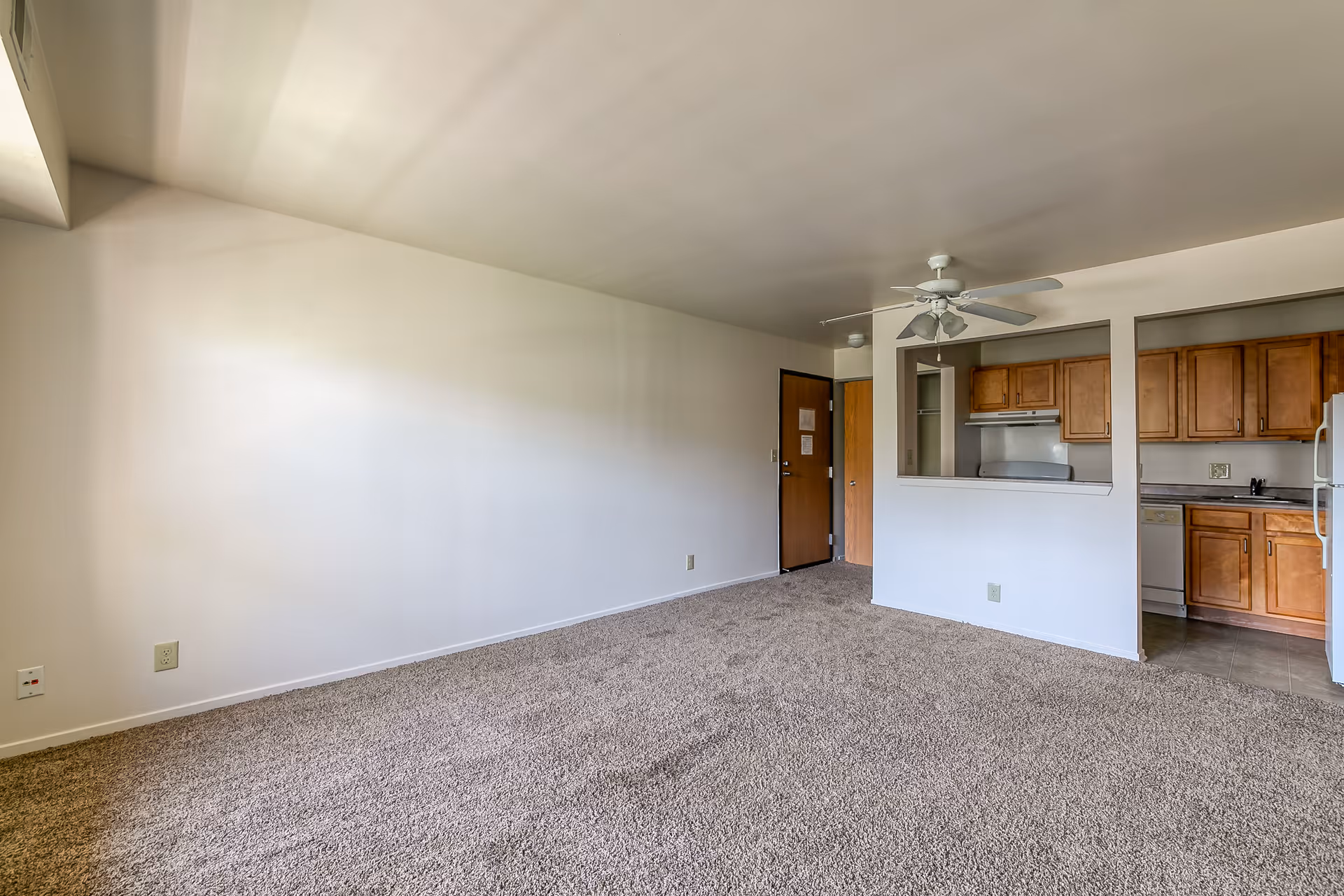 Empty room with beige carpet and white walls, featuring a ceiling fan with lights, a wooden door, and an open kitchen area with wooden cabinets and white appliances.