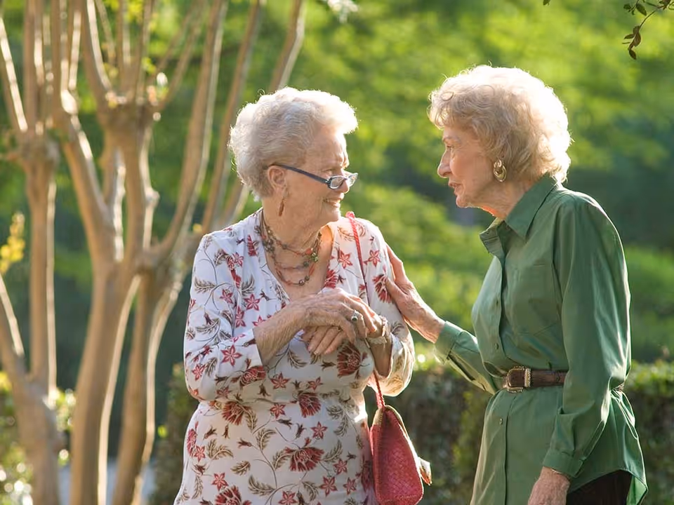 Two elderly women standing outdoors in a garden or park area, engaged in a friendly conversation. One woman is wearing a white floral blouse and glasses, holding a pink purse, while the other woman is dressed in a green shirt with a belt. Trees and greenery are visible in the background.