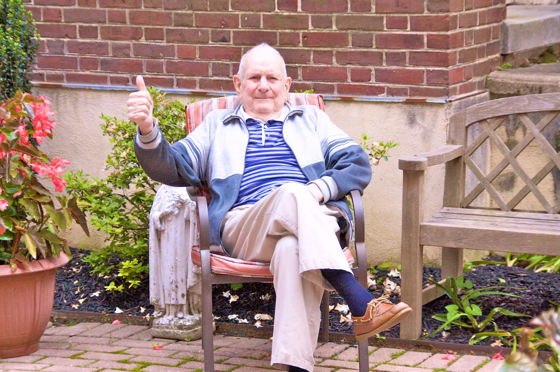An elderly man sitting on a cushioned outdoor chair in a garden area, giving a thumbs-up gesture. He is wearing a striped shirt, light jacket, beige pants, and brown shoes. Surrounding him are plants, a potted flower, a wooden bench, and a brick wall in the background.