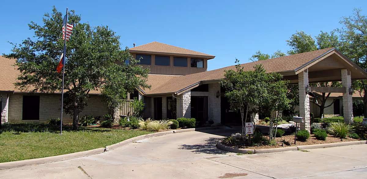 Exterior view of College Place Assisted Living facility showing a single-story building with a covered entrance, stone facade, and a driveway. There are trees and shrubs around the building, and two flagpoles with the American flag and another flag. The sky is clear and blue.