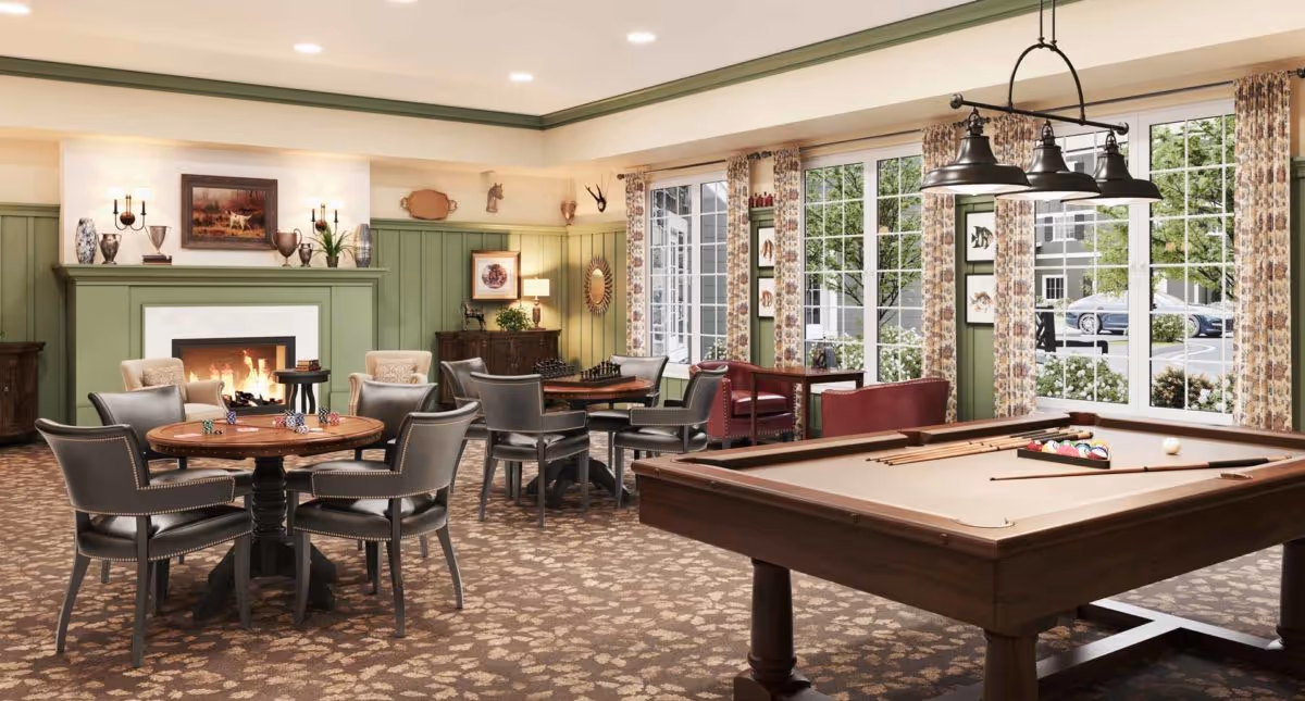 A bright communal game room with a pool table, card tables, a fireplace, and seating beside large windows.